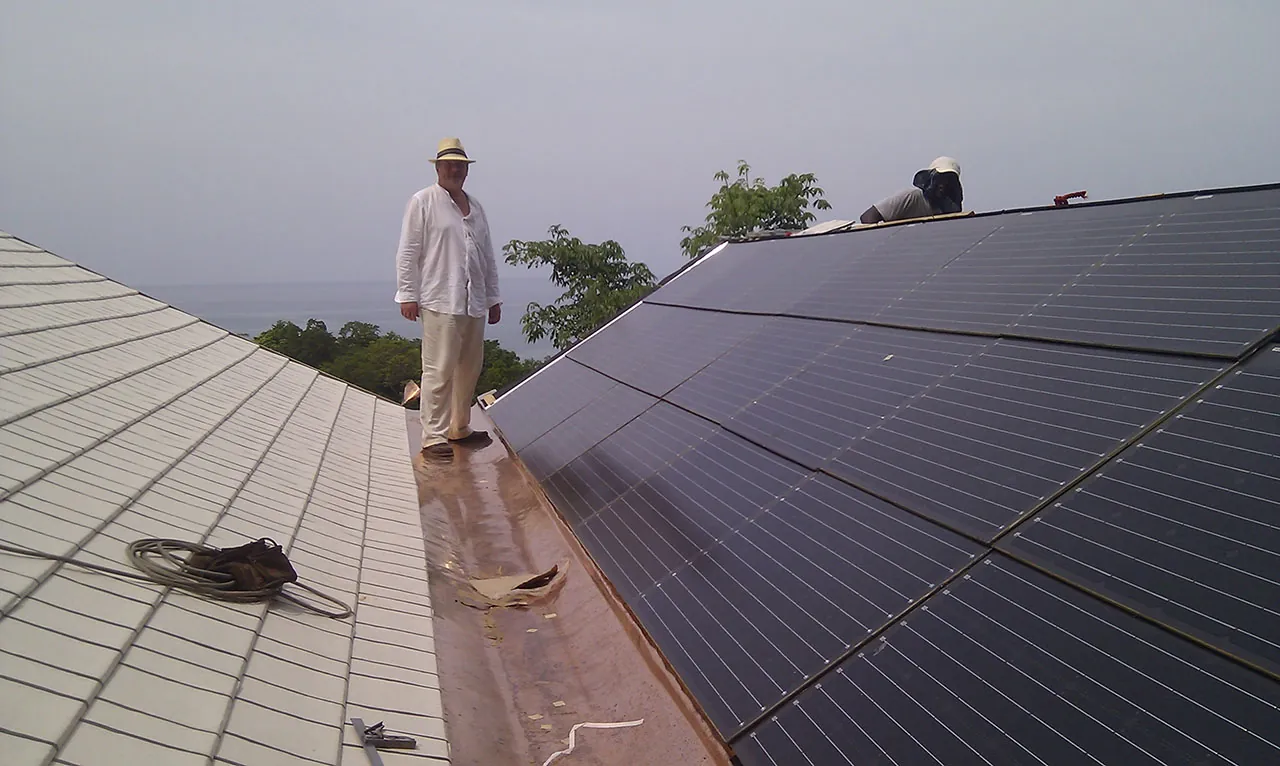 Dark solar PV panels being fitted along a roof, with trees and the sea beyond